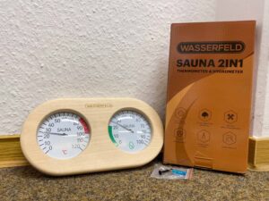 Saunathermometer und Verpackung samt Schraube und Mini-Schraubendreher auf dem Boden.