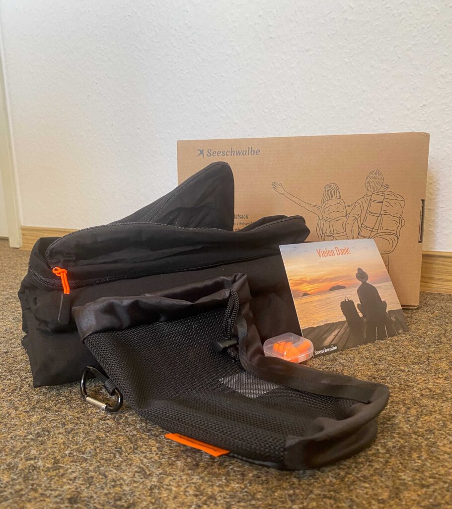 Hüttenschlafsack, Beutel, Ohrstöpsel, Dankeskarte und Verpackung aller zusammen neben- und hintereinander aufgestellt.